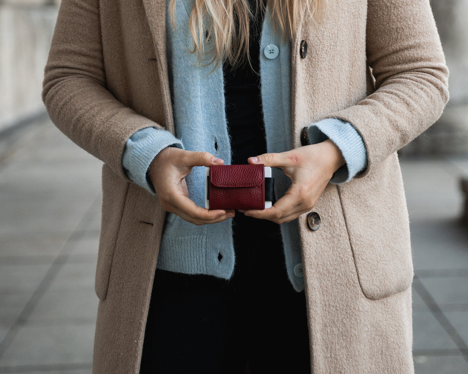 Blonde Frau mit beigem Mantel hält den Nano Boy mit roter Münztasche vor dem Körper.